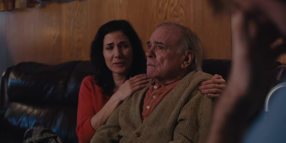 An elderly man with tears in his eyes is sad and sitting on a sofa with a young woman next to him comforting him and there is another person in front with his hand on his head in worried tones