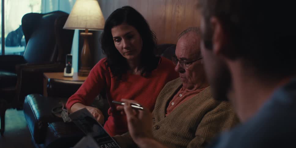 A young man is pointing with a pen at a paper that an elderly man is holding while sitting next to a woman