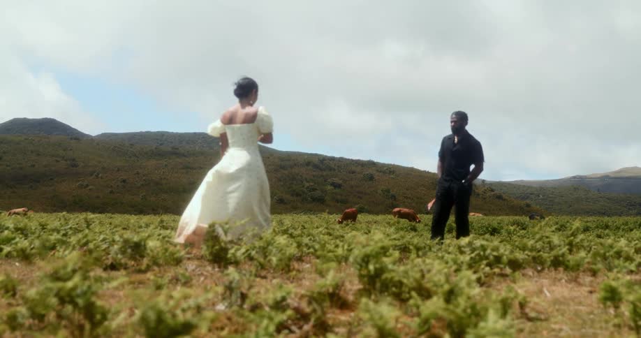 Woman in white dress walking towards a man in black outfit while they are in a field with mountains in the background
