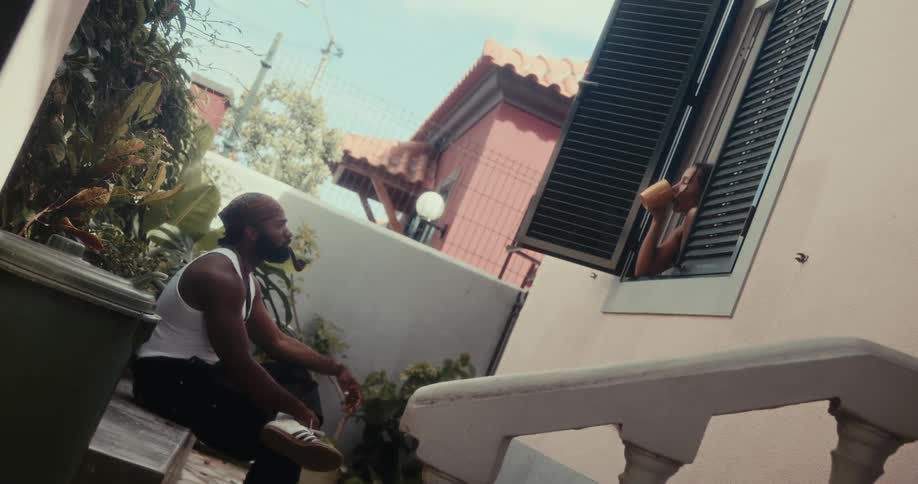 Man smoking a pipe in the backyard of the house while a woman in the window is drinking from a mug