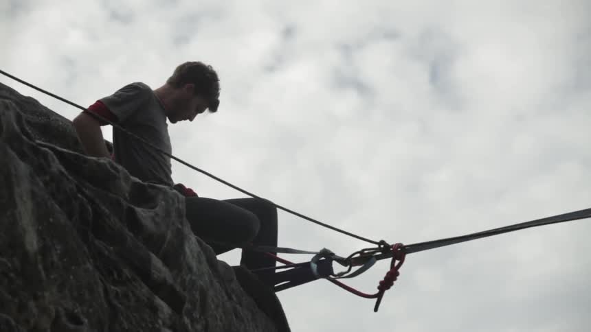 A man sits on a mountain side hooking up a large wire and harness