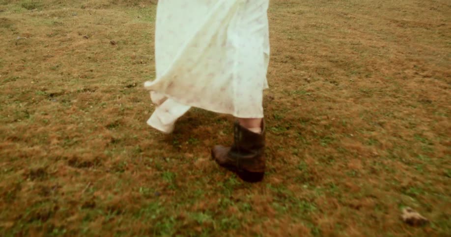 Legs of a woman in leather boots and a long white dress walking on the grass