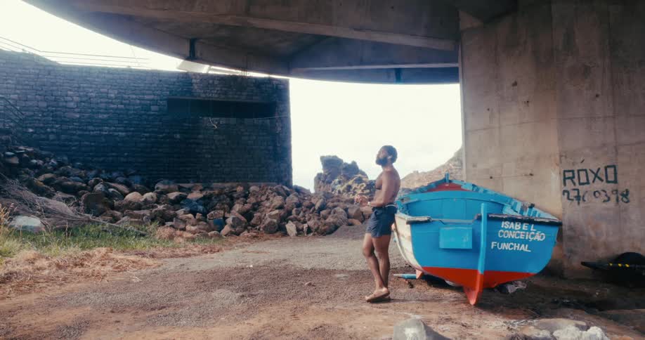 A person stands next to blue and red boat sits on a dirt and rock surface under a bridge