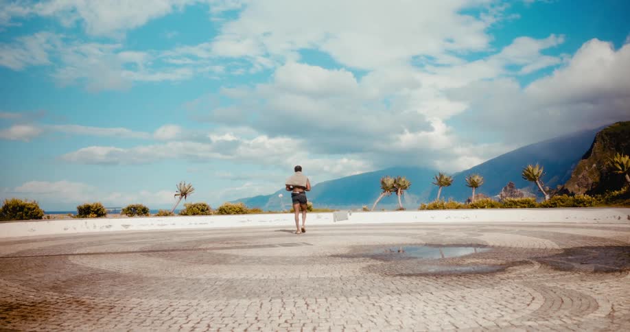 A black man walks across a concrete court near a tropical ocean coast on a partly cloudy day
