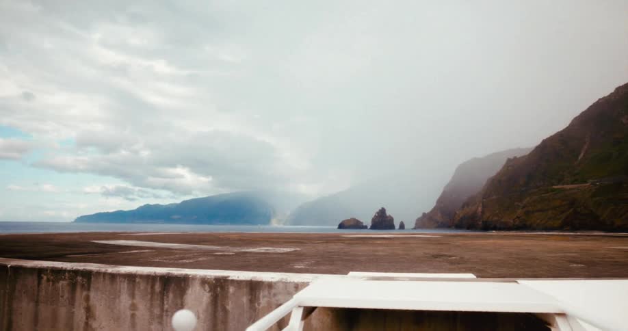 A concrete helipad extending over a rocky cliffside with a vast ocean backdrop under a cloudy sky and distant mountains fading into the horizon