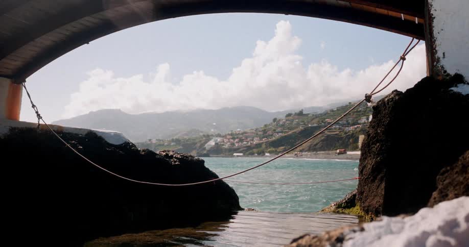 A curved concrete bridge frames a distant coastal scene with a town nestled between rolling hills and a blue sky with fluffy white clouds