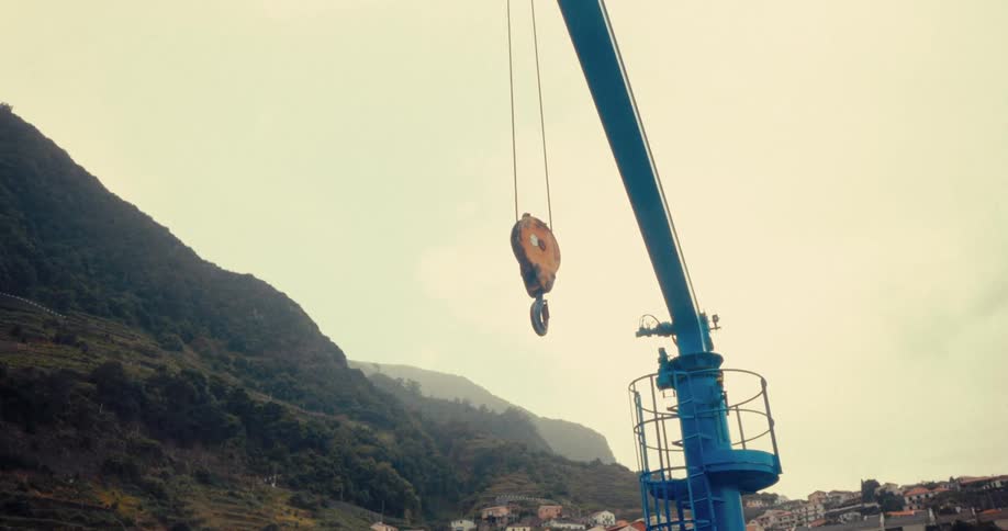 A blue crane hook hangs suspended against a backdrop of hazy hills and a cloudy sky