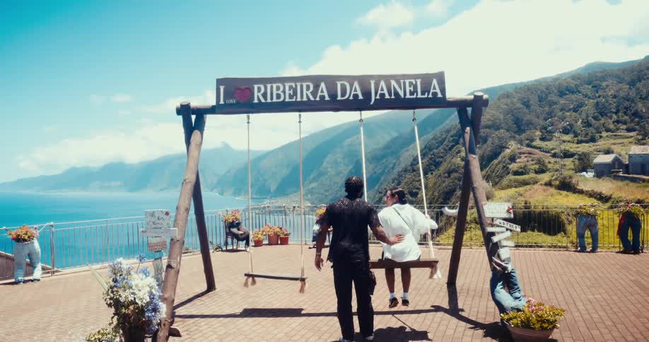 Man standing pushing a woman on a swing located on a lookout overlooking the sea and green mountains