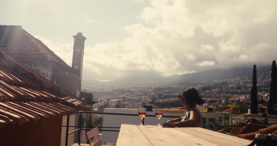 A woman is sitting at a wooden table on a rooftop terrace with two glasses of wine overlooking a cityscape with mountains in the background under a partly cloudy sky