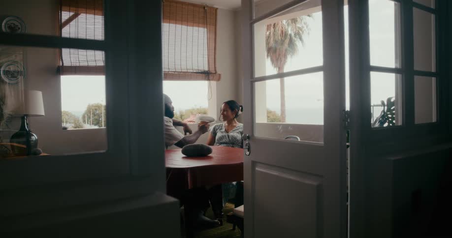 A couple sharing a drink from a cup while seated in a room with windows overlooking palm trees and the ocean