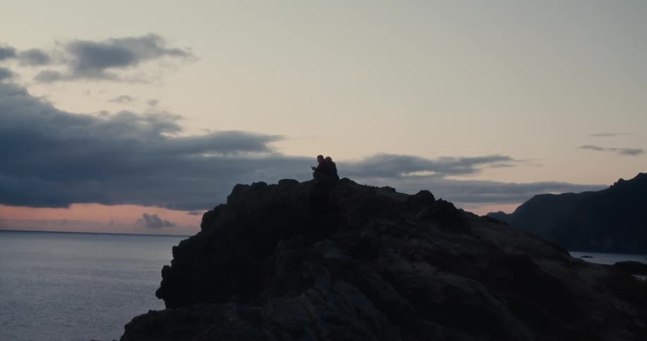 Silhouette of two people sitting on a rocky shore by the sea at dawn