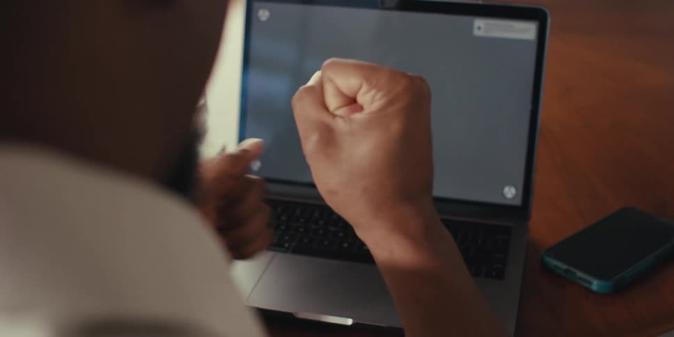 The hands of a man are shown celebrating and clapping in front of a laptop with a grey screen