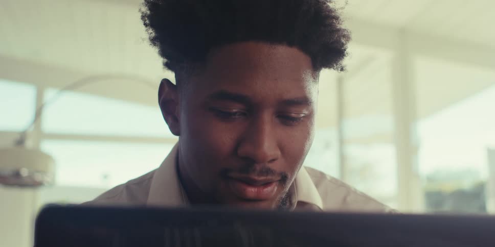 A black man sits indoors and contemplates while working on his laptop