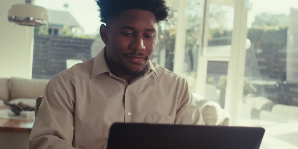 A black man is sitting at a wooden table and video conferencing on his laptop