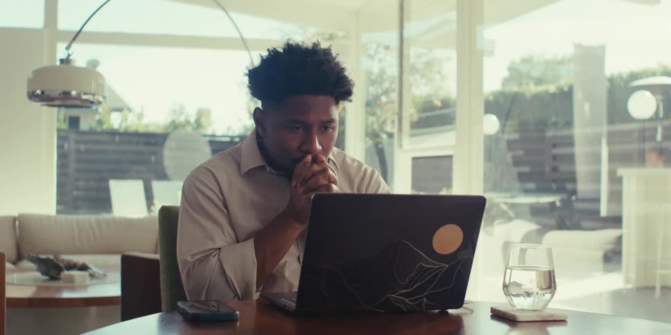 A man in a brown shirt sits impatiently at a table in front of his laptop