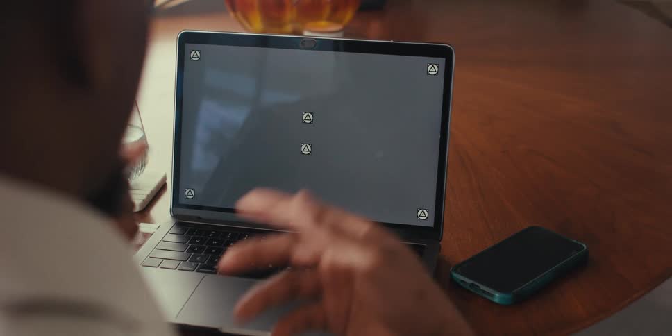 A person is gesturing with both hands in front of a laptop on a wooden table with a smartphone placed nearby