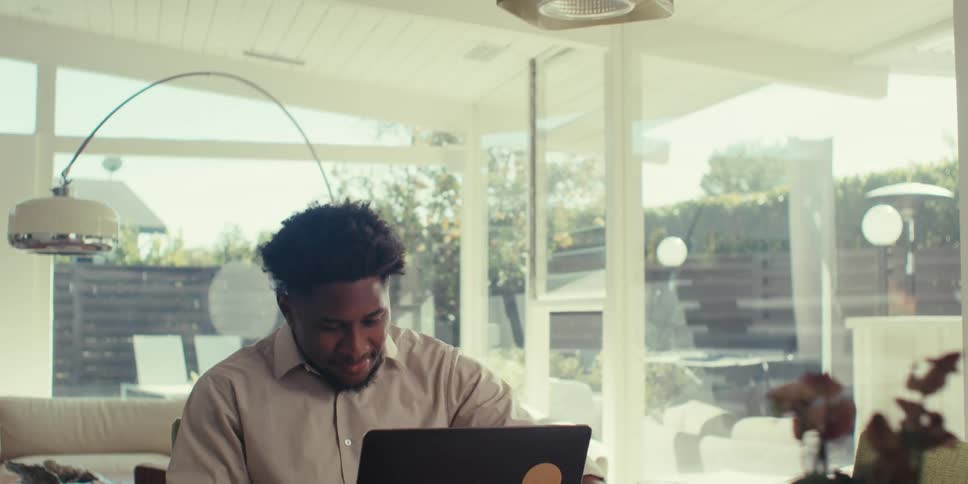 A person is sitting at a table in a modern welllit room drinking from a cup while working on a laptop and celebrating