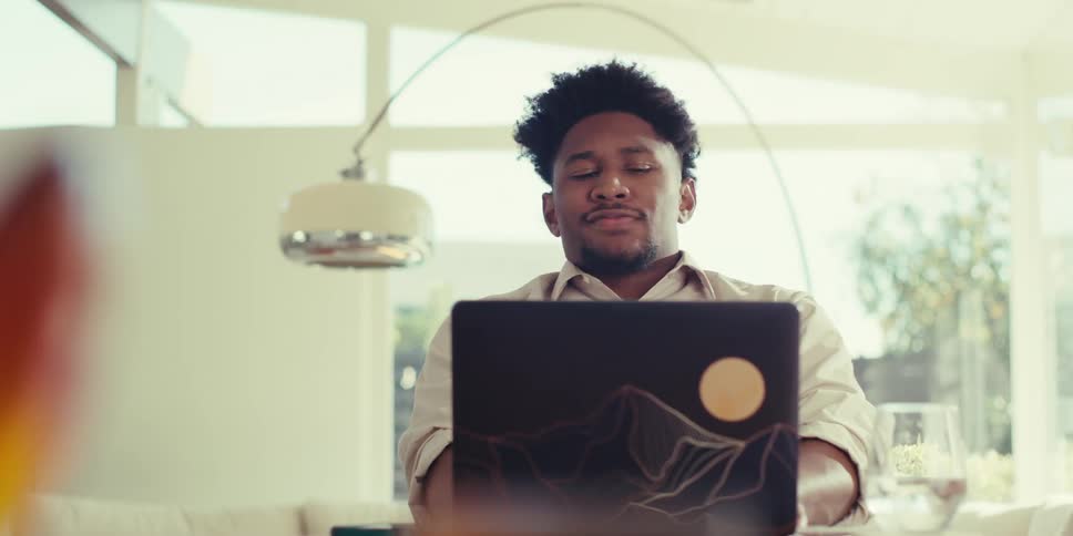 A pensive man in a brown shirt sits at a table in his living room at home using his laptop before pausing to pick up his mobile phone and leave