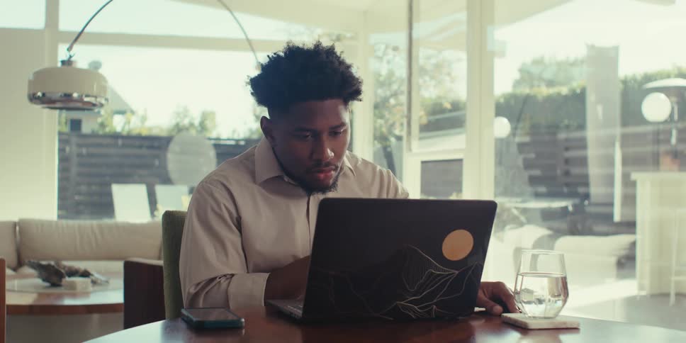 A person is sitting at a table in a bright modern room with large windows gesturing while working on a laptop