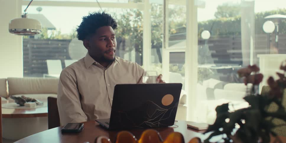 A person is sitting at a table in a modern welllit room drinking from a cup while working on a laptop