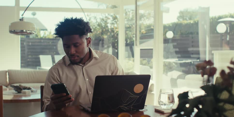 Man wearing a brown shirt sits at table using a laptop and a smartphone in the living room at home