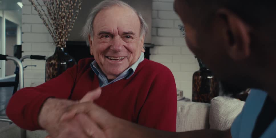 An elderly person sitting next to a walker shakes hands with a caregiver in a modern spacious living room with