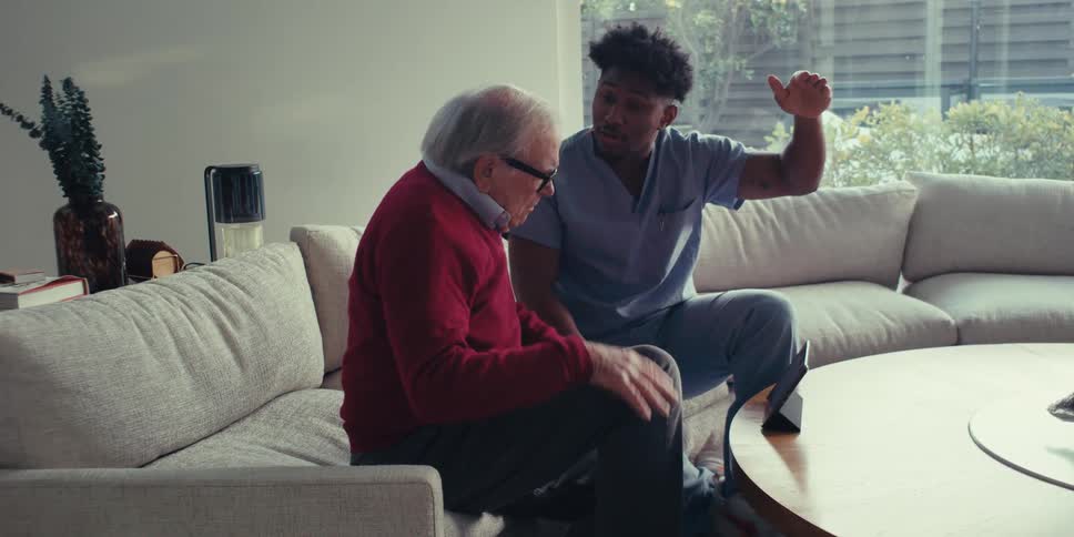 An elderly man in a red sweater and glasses talks with a healthcare worker in blue scrubs who is sitting on a beige couch