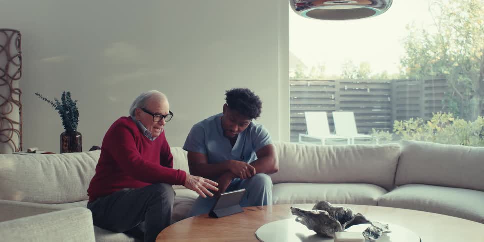 An elderly man and his caregiver sit on a couch on a spacious modern room using a tablet