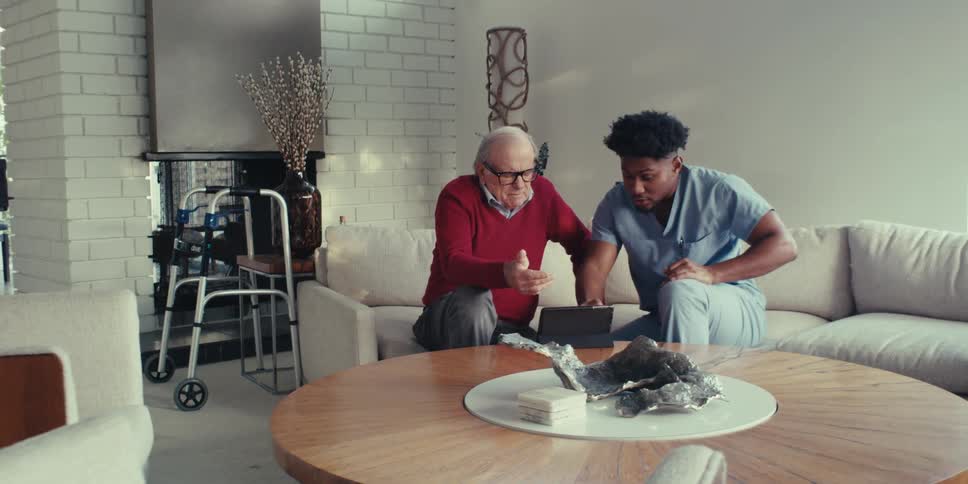 Nurse monitoring an elderly patient while they both sit on a couch interacting in front of a tablet