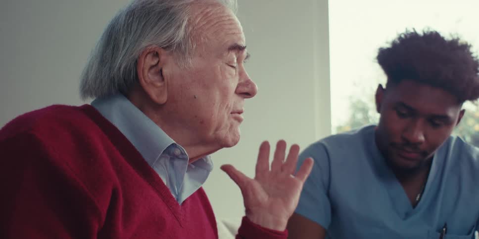 An elderly man in a red sweater gesturing with his hand while communicating with a healthcare worker in blue scrubs