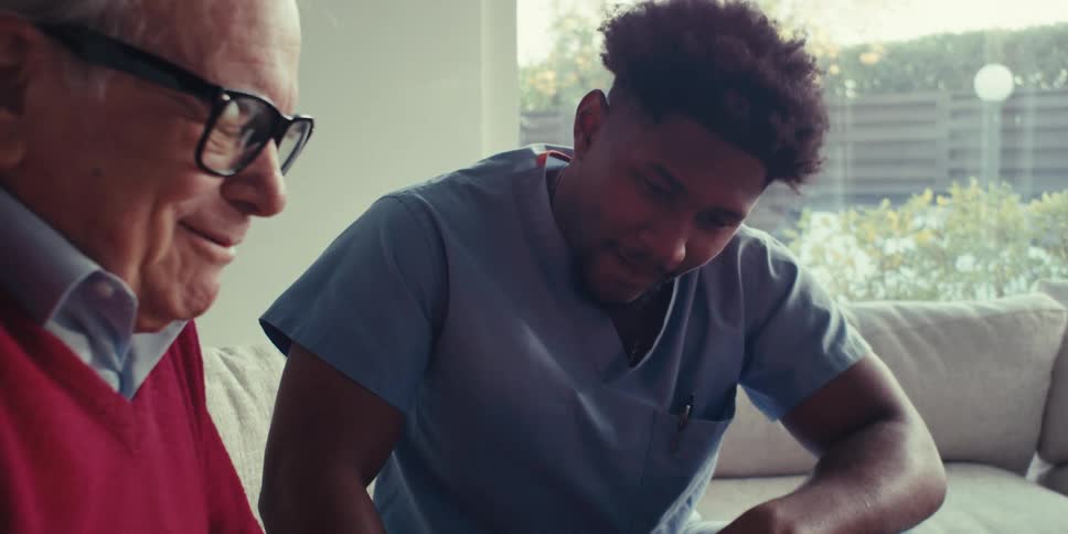 Home care professional and an elderly man smile while talking in the living room on a sunny day