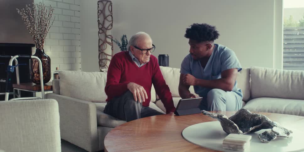 Nurse sitting next to an elderly patient and both interacting with a tablet in front of them