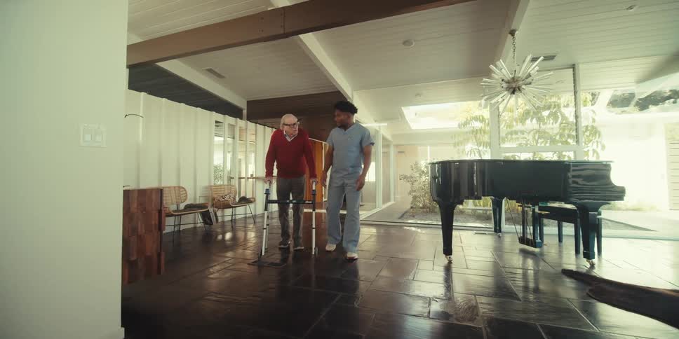 An elderly person using a walker is being assisted by a caregiver in a modern spacious living room with a grand piano and large windows