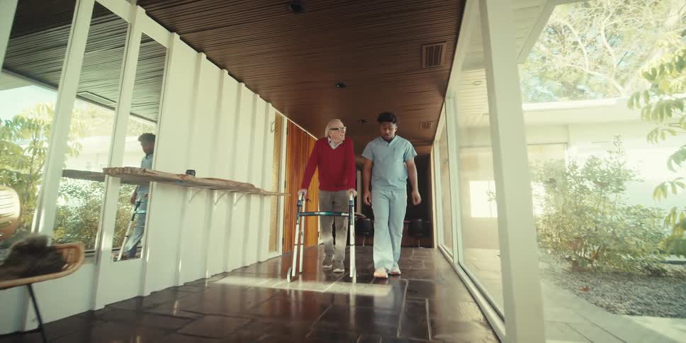 Nurse talking to elderly man who bursts out laughing as he walks down a hallway