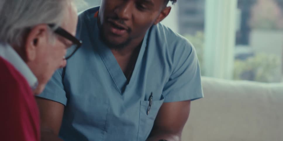 An elderly man in a red sweater and glasses gestures with his hand while communicating with a healthcare worker in blue scrubs who is sitting on a beige couch