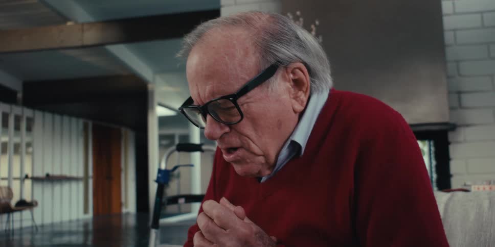 An elderly person sits on a couch next to their walker reading from a tablet held by another person sitting beside him