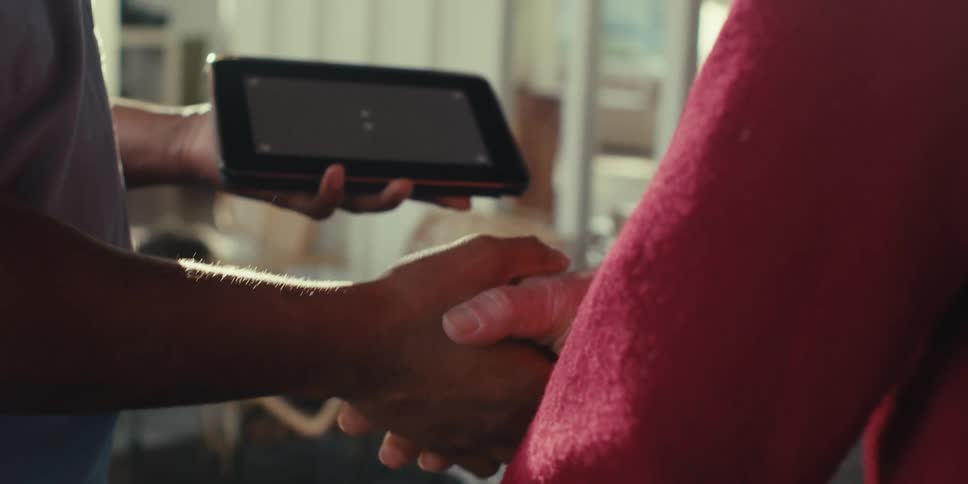 Two people agree with a handshake one of them holding a tablet with a gray screen in a brightly sunlit room