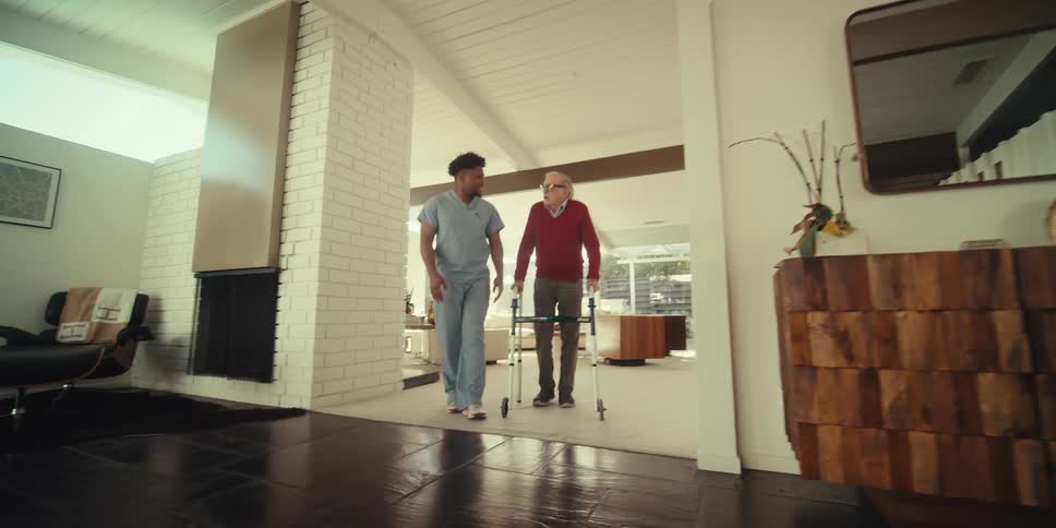 An elderly person using a walker is being assisted by a caregiver in a modern spacious living room with a grand piano and large windows