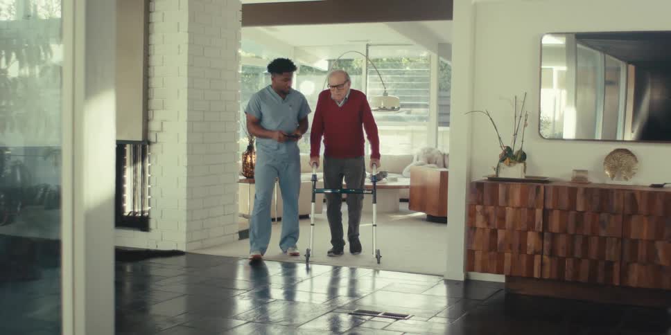 An elderly person using a walker is being assisted by a caregiver in a modern spacious living room with large windows