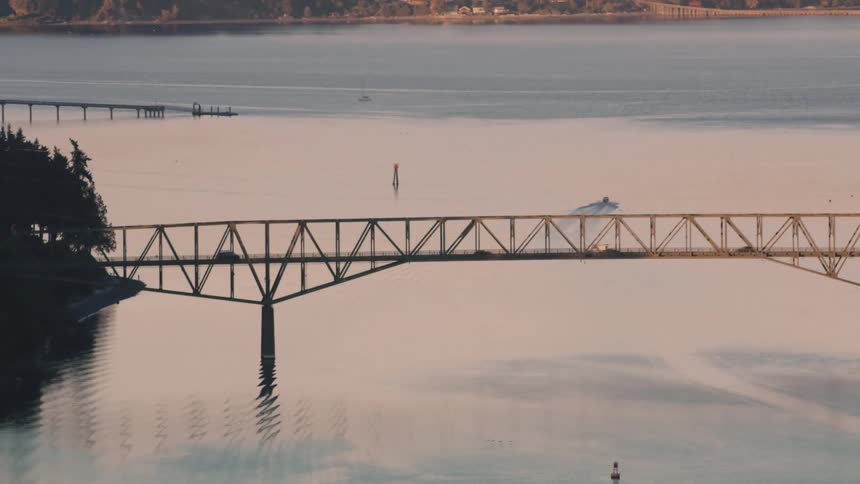 Vehicles cross a bridge across the water from houses nestled in the woods on a foggy day