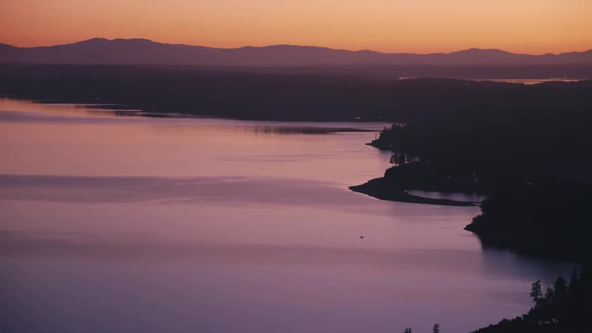 A still lavendertinted lake reflects the silhouette of distant mountains against a vibrant sunset sky