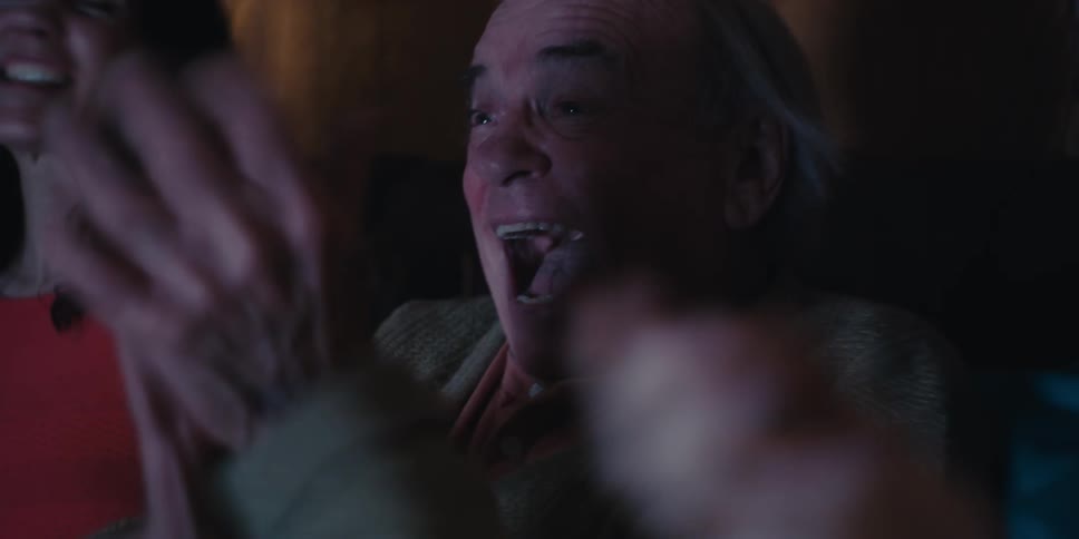 Elderly man sitting in the middle of two young men and they all smiling and cheering as they sit on the living room couch