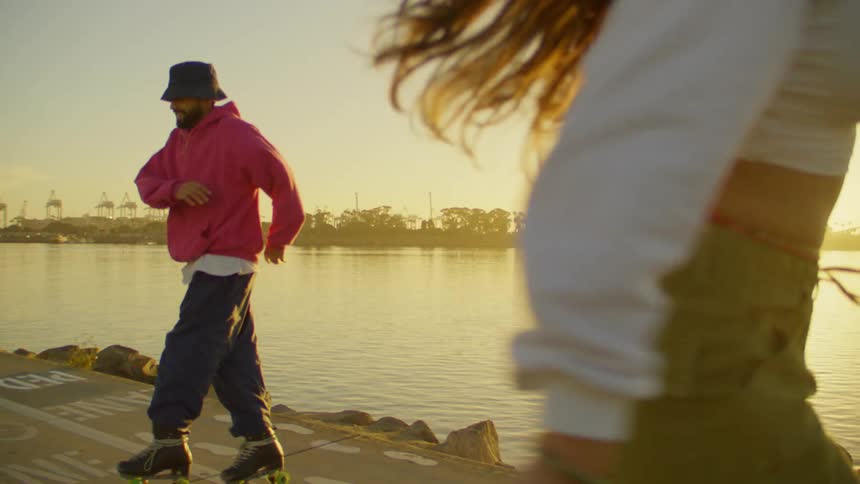 Man and woman in the foreground dancing on roller skates by a lake