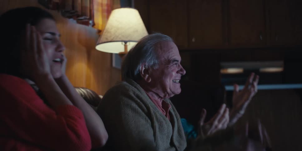Elderly man sitting next to two young adults watching television and cheering
