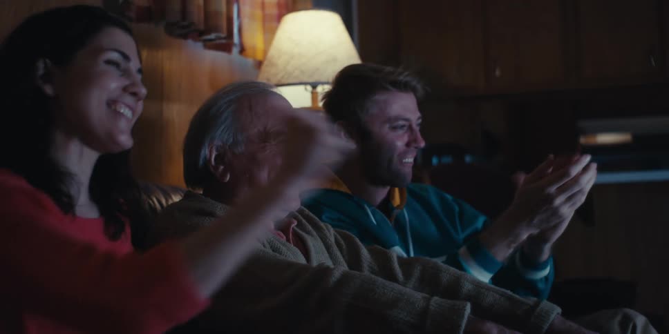 A family of three is sitting on a couch in a dimly lit room watching television together and celebrating