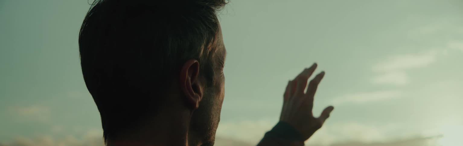 A man with his hand raised looks at the setting sun under a clear blue sky