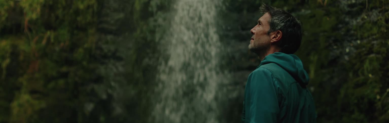 A person in a green hoodie is standing near a waterfall looking up at the cascading water