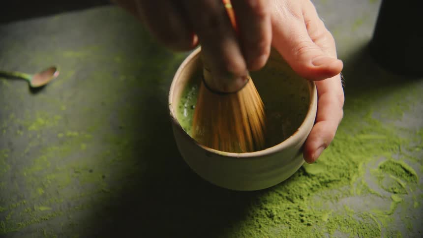A hand is using a bamboo whisk to mix green matcha powder in a bowl