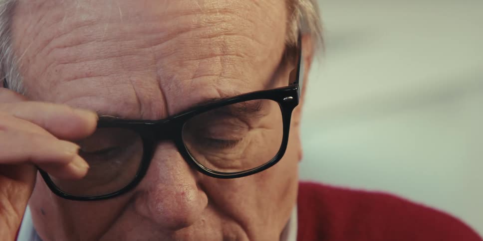 An elderly person wearing glasses and a red sweater looks intently at something offcamera
