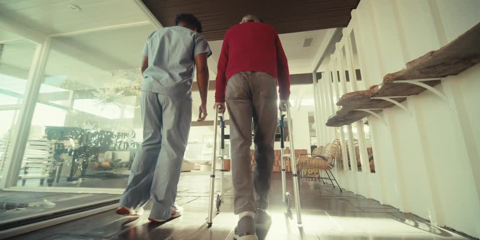 Man accompanying an elderly man who walks with the help of a walker down a hallway